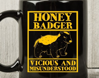 May include: Black ceramic mug with a yellow and black honey badger graphic. The mug features the text "HONEY BADGER" above an illustration of the animal, and "VICIOUS AND MISUNDERSTOOD" below. The mug has a large, curved handle.