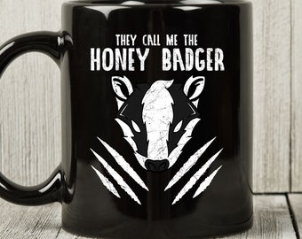May include: Black ceramic mug with a white graphic of a honey badger and the text "THEY CALL ME THE HONEY BADGER". The mug features a black handle and a white claw mark design.