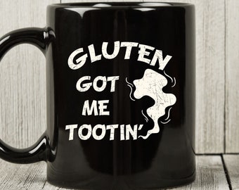 May include: Black ceramic mug with the text "GLUTEN GOT ME TOOTIN'" in white. The mug has a curved handle and a humorous design, perfect for coffee or tea.