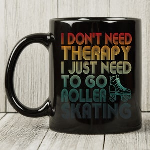Puede incluir: Taza de cerámica negra con el texto "I DON'T NEED THERAPY I JUST NEED TO GO ROLLER SKATING" en un esquema de colores retro. La taza presenta un gráfico de patines. La taza tiene un asa grande y está sobre un fondo de madera clara.