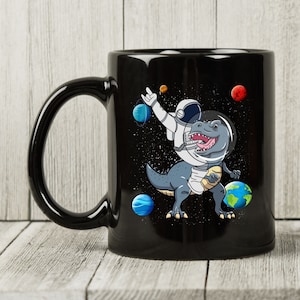 May include: Black ceramic mug featuring a cartoon dinosaur in an astronaut helmet and suit, dabbing in space. Planets and stars surround the dinosaur. The mug has a large, curved handle.