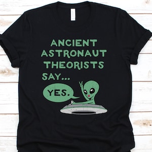 May include: Black t-shirt with a green graphic of an alien in a spaceship. The text reads "ANCIENT ASTRONAUT THEORISTS SAY... YES." The alien is giving a peace sign. The shirt is made of a soft material.