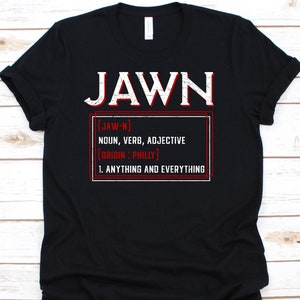 以下が含まれることがあります： 黒いTシャツに白い文字で「JAWN」と書かれています。その下に、赤枠で「JAWN」を名詞、動詞、形容詞として定義し、フィラデルフィア発祥で「すべて」を意味すると説明しています。