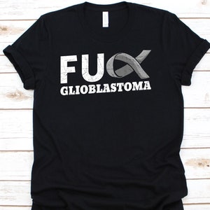 May include: Black t-shirt featuring the words "FUCK GLIOBLASTOMA" in a distressed white font. A grey awareness ribbon is part of the design. The t-shirt is made of a soft material.