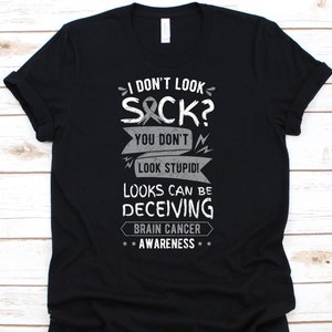 May include: Black t-shirt with white and grey text. The text reads "I DON'T LOOK SICK? YOU DON'T LOOK STUPID! LOOKS CAN BE DECEIVING BRAIN CANCER AWARENESS". The shirt has a crew neck and short sleeves.