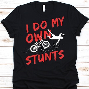 May include: Black t-shirt with the text "I DO MY OWN STUNTS" in red, along with a white graphic of a BMX bike and a person performing a stunt. The design is printed on the front of the shirt.