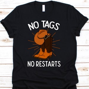 以下が含まれることがあります： 黒いTシャツに白い文字で「NO TAGS NO RESTARTS」と書かれています。茶色のカウボーイハットとブーツ、オレンジ色の光線が描かれています。柔らかい素材で作られたシャツです。