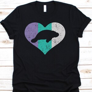 May include: Black t-shirt with a distressed heart design and a manatee silhouette. The heart is split into purple, white, and teal vertical stripes. The manatee is a solid black colour.