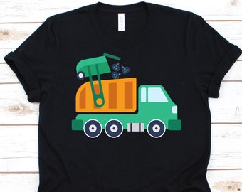 May include: Black t-shirt featuring a colorful graphic of a green and orange garbage truck. The truck is depicted with a raised container, and the design includes blue and white accents. The shirt is made of a soft material.