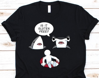 May include: Black t-shirt with a humorous graphic featuring two cartoon sharks and a person in a red and white inner tube. A speech bubble above the sharks reads "IS IT GLUTEN FREE?"