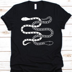 May include: Black t-shirt featuring a white snake design. The snake is stylized with crescent moon and arrow patterns along its body. The design is centered on the shirt, creating a bold graphic statement. The shirt has a classic crew neck and short sleeves.