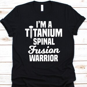 Puede incluir: Camiseta negra con texto blanco y un gráfico de tornillo. El texto dice "I'M A TITANIUM SPINAL FUSION WARRIOR". La camiseta está hecha de un material suave y tiene cuello redondo.