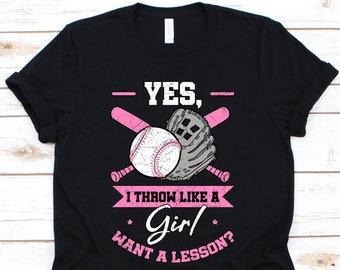 Op de afbeelding: Zwart T-shirt met een honkbalontwerp. Het ontwerp bevat een honkbal, handschoen en knuppels in roze en wit, met de tekst "YES, I THROW LIKE A Girl WANT A LESSON?"
