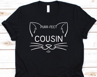 May include: Black t-shirt with a white cat face design. The design includes the words "PURR-FECT COUSIN" in a playful font, with cat ears and whiskers. A casual, comfortable tee for cat lovers.