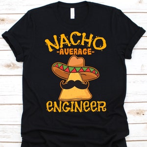 May include: Black t-shirt with a graphic of a nacho chip wearing a sombrero and mustache. The text reads "Nacho Average Engineer" in yellow and orange. The shirt is made of a soft material.