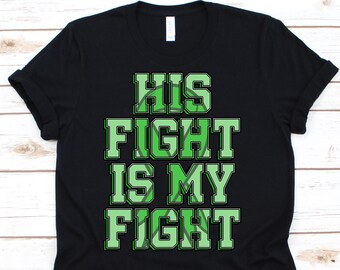 Peut inclure: T-shirt noir avec l'inscription "HIS FIGHT IS MY FIGHT" en lettres vertes. Le texte a un contour vert et un remplissage vert clair. Le t-shirt est en matière souple et convient au quotidien.