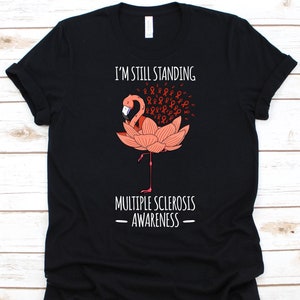 以下が含まれることがあります： 黒いTシャツ。フラミンゴのデザインと「I'M STILL STANDING MULTIPLE SCLEROSIS AWARENESS」の文字。フラミンゴはピンク色で、ピンク色の蓮の花の上に立っています。赤い啓発リボンが付いています。