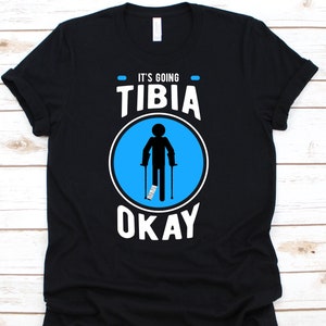 以下が含まれることがあります： 「IT'S GOING TIBIA OKAY」の文字が入った黒いTシャツ。青い円の中に、ギプスと松葉杖を持った棒人間が描かれています。カジュアルウェアです。