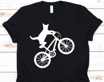 May include: Black t-shirt featuring a white graphic of a cat riding a bicycle. The cat is depicted in a playful pose, with its front paws on the handlebars. The bicycle is also white, creating a striking contrast against the black fabric.