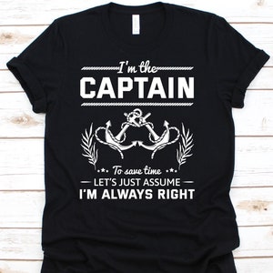 Op de afbeelding: Zwart T-shirt met witte tekst: "I'm the CAPTAIN." Daaronder een afbeelding van twee gekruiste ankers met een bladmotief. De tekst eronder luidt: "To save time... LET'S JUST ASSUME I'M ALWAYS RIGHT."