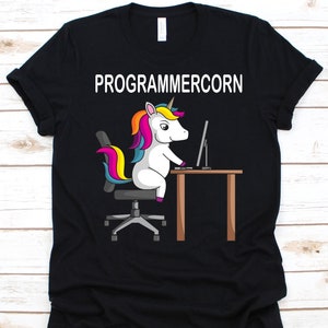 May include: Black t-shirt with the word "PROGRAMMERCORN" above a cartoon unicorn sitting at a desk. The unicorn has a rainbow mane and tail and is facing a computer monitor. The desk is brown.