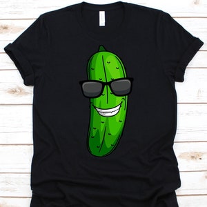 Może przedstawiać: Czarny t-shirt z kreskówkową ilustracją zielonego ogórka w okularach przeciwsłonecznych i uśmiechem. Ogórek ma biały uśmiech i czarne okulary przeciwsłoneczne. Koszulka wykonana jest z miękkiego materiału.