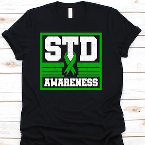 May include: Black t-shirt with a green and white graphic design. The design includes the text "STD AWARENESS" in white block letters, a green ribbon, and a green border. The shirt is made of a soft material.