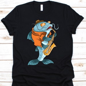 Puede incluir: Camiseta negra con un dibujo de un pez gato de dibujos animados tocando el saxofón. El pez gato es azul con ropa naranja, gafas de sol y un sombrero marrón. El saxofón es dorado y marrón.