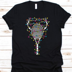 May include: Black t-shirt featuring a white tennis racket design wrapped in colorful Christmas lights. The racket's handle and frame are white, and the lights are various colors. A festive graphic tee for tennis enthusiasts.