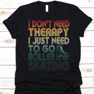 Puede incluir: Camiseta negra con el texto "I don't need therapy, I just need to go roller skating" en una fuente retro. Un gráfico de patines de ruedas turquesa aparece debajo del texto. La camiseta es de cuello redondo clásico.