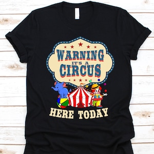 Puede incluir: Camiseta negra con un gráfico colorido con temática de circo. El diseño presenta una carpa de circo, un payaso malabarista y un elefante azul equilibrándose sobre una pelota. El texto dice "WARNING IT'S A CIRCUS HERE TODAY."