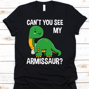 以下が含まれることがあります： 黒いTシャツ。緑色の恐竜が腕を負傷し、「CAN'T YOU SEE MY ARMISSAUR?」の文字。恐竜は黄色い腕と白い包帯をしています。Tシャツはクルーネックです。