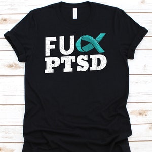 May include: Black t-shirt with the text "FUCK PTSD" in white, distressed font. A teal ribbon graphic is incorporated into the design. The shirt is made of a soft material and has a crew neck.