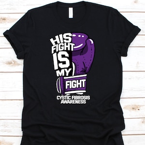 Puede incluir: Camiseta negra con un diseño gráfico que presenta un guante de boxeo morado y el texto "HIS FIGHT IS MY FIGHT" en blanco. Debajo del texto está "CYSTIC FIBROSIS AWARENESS" en blanco.