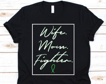 May include: Black t-shirt with the words "Wife. Mom. Fighter." in a white box, with green accents and a green ribbon. The shirt is made of a soft material and has a classic crew neck.