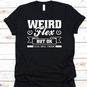 Puede incluir: Camiseta negra con texto blanco: "WEIRD Flex BUT OK" encima de "EHLERS DANLOS SYNDROME". El texto es en negrita, sin serifa. La camiseta está doblada, mostrando el frente de la prenda.