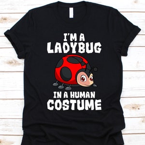May include: Black t-shirt with the text "I'M A LADYBUG IN A HUMAN COSTUME" in white. A cartoon ladybug with a red shell and black spots is centered below the text. The ladybug has a smiling face.