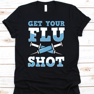 Könnte beinhalten: Schwarzes T-Shirt mit dem Text "GET YOUR FLU SHOT" in Weiß und Hellblau. Zwei hellblaue Spritzen sind unter dem Text gekreuzt. Das Shirt hat einen Rundhalsausschnitt.