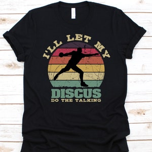 Może przedstawiać: Czarna koszulka z retro wzorem. Przedstawia sylwetkę dyskobola na tle zachodu słońca. Tekst brzmi "I'll let my discus do the talking" w stylu vintage, idealna dla fanów sportu.