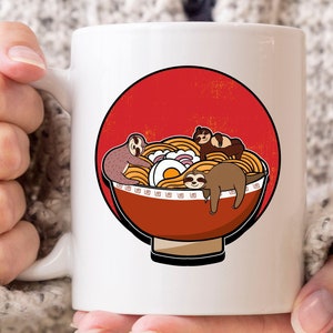 May include: White ceramic mug featuring a cartoon illustration of sloths in a bowl of ramen noodles. The bowl is red with a black outline, set against a red circle. The sloths are brown and the ramen is yellow.