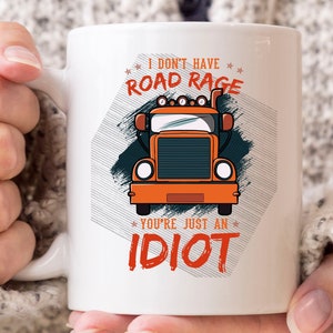 May include: White ceramic mug featuring a cartoon orange semi-truck design with the text "I don't have road rage, you're just an idiot." The mug has a large handle and is held by a person.