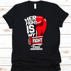 May include: Black t-shirt with a red boxing glove graphic and the text "HER FIGHT IS MY FIGHT" in white. Below the glove, the text "STROKE AWARENESS" is displayed in white. The shirt is made of a soft material.