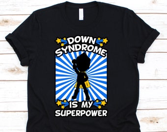 Op de afbeelding: Zwart T-shirt met de tekst "DOWN SYNDROME IS MY SUPERPOWER". Het ontwerp toont een silhouet van een kind in een superheldenpose, tegen een blauw-witte burst, met gele sterren. Een statement-T-shirt.