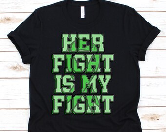 May include: Black t-shirt with the text "HER FIGHT IS MY FIGHT" in green block letters. The text has a white outline and a green design inside the letters. The shirt is folded on a light wood surface.