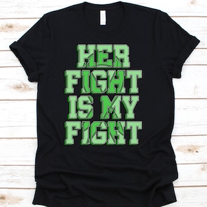 Może przedstawiać: Czarna koszulka z napisem "HER FIGHT IS MY FIGHT" w zielonych literach blokowych. Tekst ma biały kontur i zielony wzór wewnątrz liter. Koszulka jest złożona na jasnej drewnianej powierzchni.