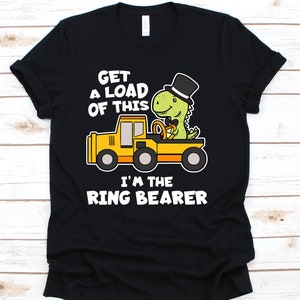 May include: Black t-shirt with a graphic of a green dinosaur wearing a top hat, driving a yellow truck. Text reads "GET A LOAD OF THIS I'M THE RING BEARER."