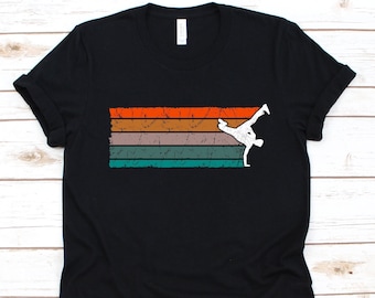 Puede incluir: Camiseta negra con un diseño retro. Presenta rayas horizontales en naranja, marrón, gris y turquesa. Una silueta blanca de un bailarín de breakdance está a la derecha. La camiseta tiene cuello redondo clásico.