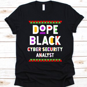 May include: Black t-shirt with the words "DOPE BLACK CYBER SECURITY ANALYST" in colorful block letters. The design includes a red, green, and yellow triangle border at the top and bottom of the text.