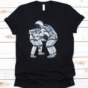 May include: Black t-shirt with a graphic of two astronauts wrestling in space. The illustration is in grey and white, with a speckled background. The t-shirt is made of a soft material.