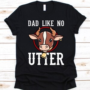 Puede incluir: Camiseta negra con el texto blanco "DAD LIKE NO UTTER". La camiseta presenta una cabeza de vaca de dibujos animados dentro de un círculo rojo. La vaca tiene pelaje marrón y blanco, cuernos y una campana.